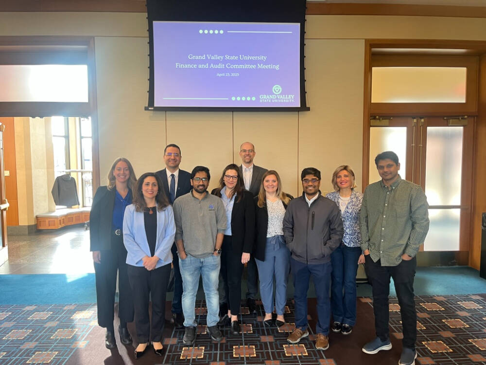 College of computing's faculty and staff stand together and smiles for a photo in front of a screen displaying “Grand Valley State University Finance and Audit Committee Meeting – April 25, 2025.”